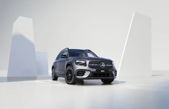 Mercedes-Benz GLB in Grau, vordere Ansicht in einem minimalistischen Studio mit weißen geometrischen Strukturen im Hintergrund.