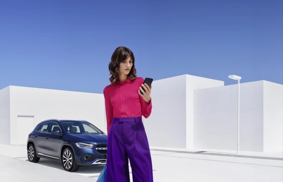 Frau in auffälligem Outfit schaut auf ihr Handy, vor einem blauen Mercedes-Benz SUV in moderner, weißer Umgebung.